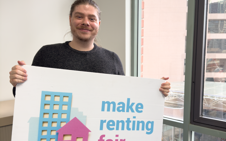 Dylan holding a sign saying make renting fair in front of a window.