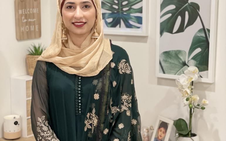 A woman smiles at the camera, standing in front of a desk and artwork on the walls. She is wearing a green dress, golden headscarf, with red lipstick and golden earrings.