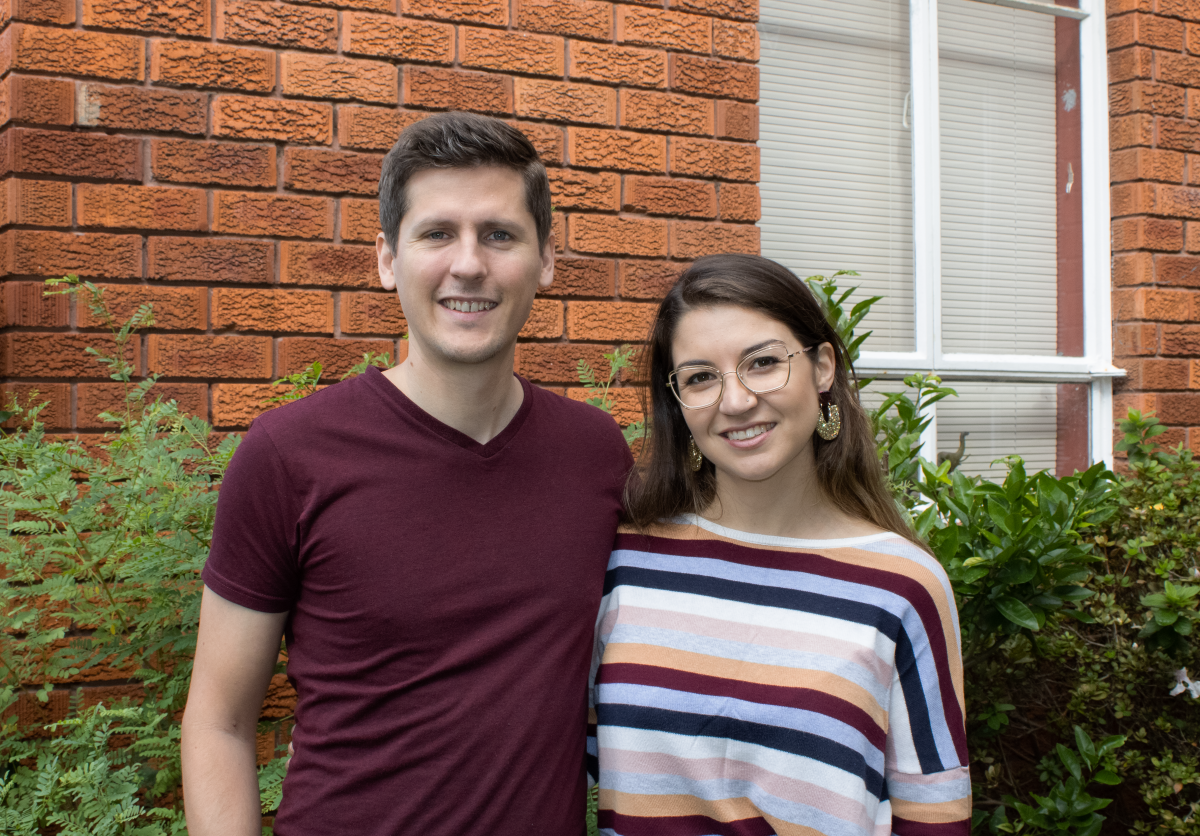 Jason and Lauren are standing close and smiling in front of a red brick wall and white window.