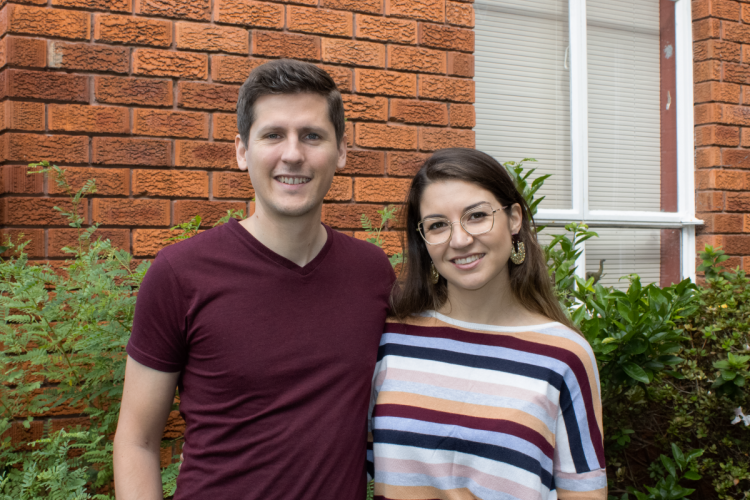 Jason and Lauren are standing close and smiling in front of a red brick wall and white window.